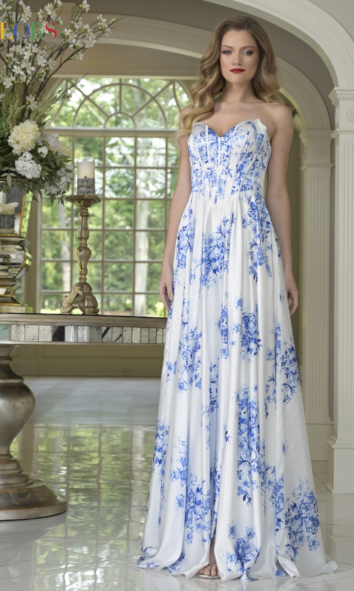 A woman poses indoors in the Strapless Blue Print Prom Dress: Colors Dress 3857, featuring elegant floral patterns. Large windows and a floral arrangement in the background enhance the beauty of this blue gown.